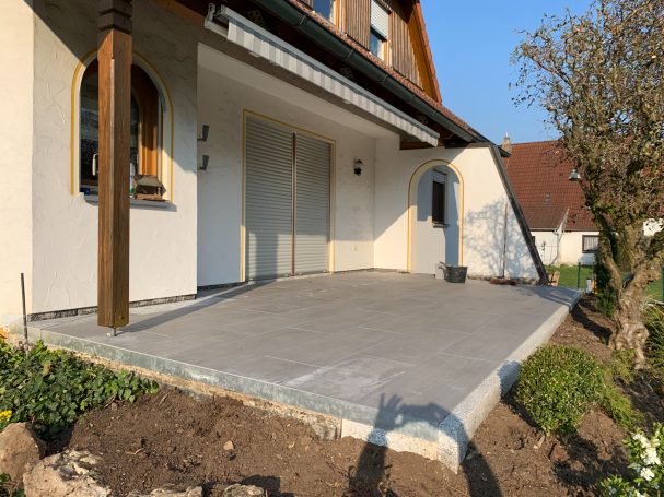 Terrasse mit neuem Plattenbelag, umgeben von Gartenpflanzen und einem hellen Himmel.