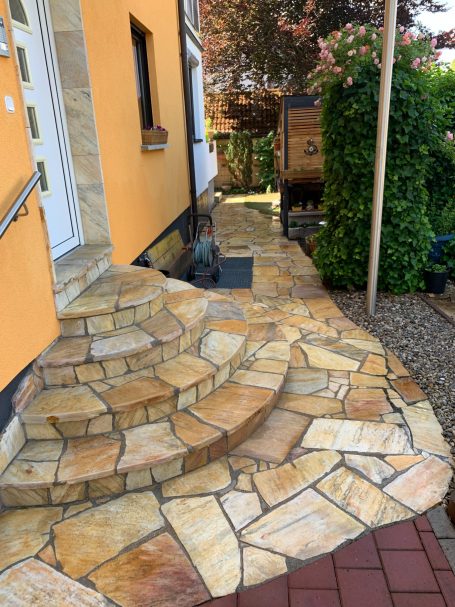 Bepflasterter Weg mit geschwungenen Stufen zu einem Haus, umgeben von Pflanzen.