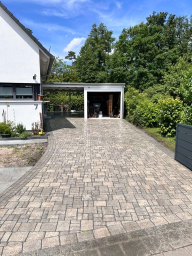 Pflastersteinerne Einfahrt mit Garage und umgeben von Bäumen und Sträuchern.