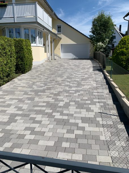 Pflastersteindriveway vor einem Wohnhaus mit umgebenden Hecken und Bäumen.