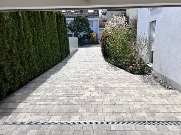 Fester Pflasterweg zwischen Hecken und Bäumen, führt zu einem Gebäude im Hintergrund.