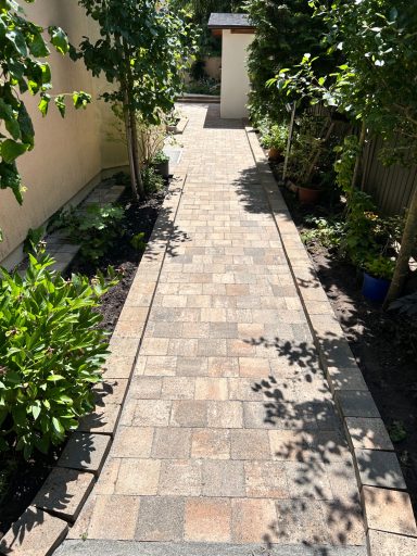 Pflasterweg zwischen Pflanzen, führt zu einem kleinen Gebäude im Hintergrund.