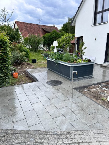 Gepflasterter Gartenweg mit Hochbeet, umgeben von Pflanzen und einem hellen Himmel.