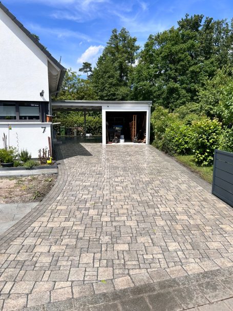 Pflastersteingartenweg mit Carport und grüner Vegetation im Hintergrund.